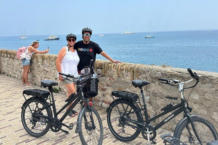 Two people on a stone wall with bicycles, a body of water, and boats in the background. 4