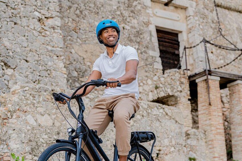 A person riding a bicycle with a helmet, in front of a stone building. 3