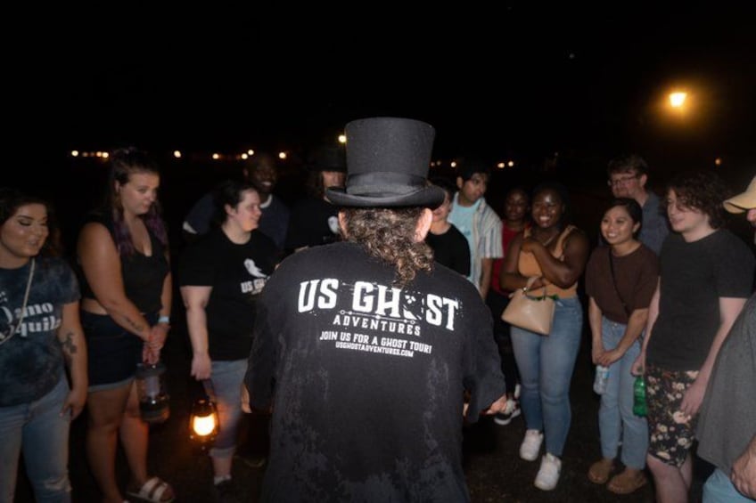 Iemand in een zwart shirt met de tekst 'US GHOST', met een zaklamp in de hand, staat 's nachts in een groep. 3