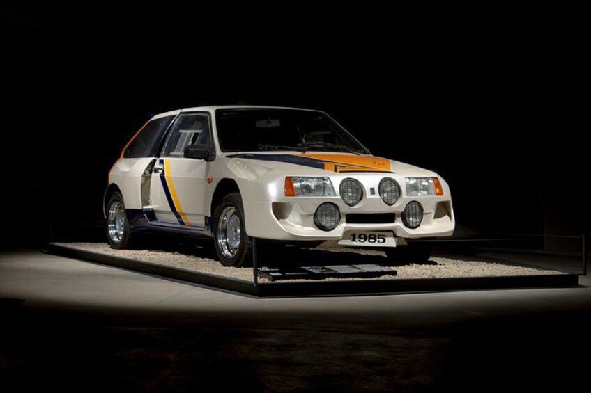 Ein weißer Rallyewagen mit gelben und blauen Streifen, ein Kennzeichen mit der Aufschrift '1985' und einem Heckspoiler. 3