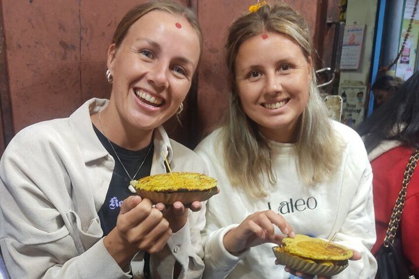 Two individuals holding bowls of food, likely a traditional dish, in a casual setting with a wooden background. 2