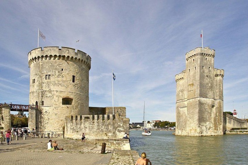 A historic stone castle with two towers, a river in the foreground, and people walking along the riverside. 3