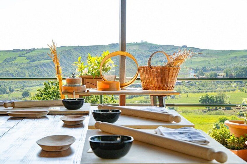 A rustic kitchen setup with rolling pins, bowls, and a basket on a wooden table, overlooking a green valley. 1