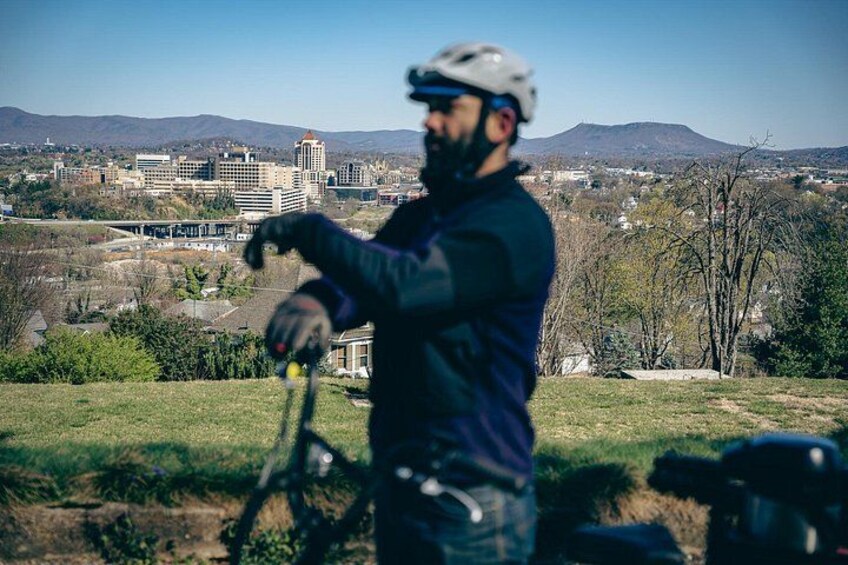 Eine Person mit Kamera auf Stativ, mit Helm, steht auf einer grasbewachsenen Anhöhe mit Stadtansicht im Hintergrund. 2