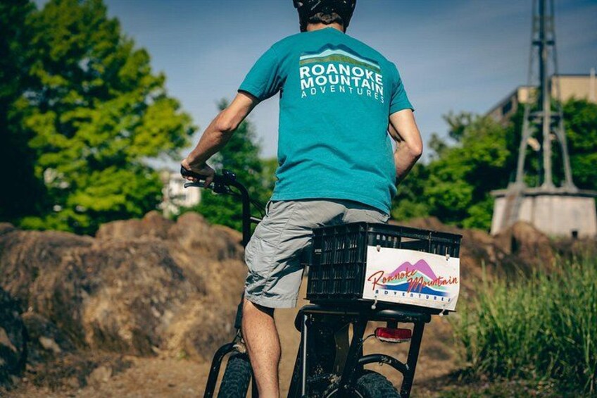 Eine Person fährt mit einem Fahrrad mit Korb, trägt ein türkisfarbenes Hemd und graue Shorts, im Hintergrund eine felsige Landschaft und Grünflächen. 5