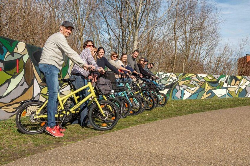 Un grupo de personas en bicicletas, alineadas frente a un muro con un mural colorido. 3