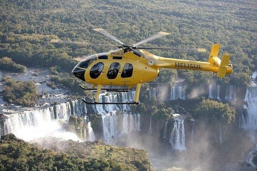 A yellow helicopter with 'PR-HLS' and 'HELISUL' markings flying over a waterfall. 1