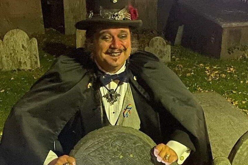 Una persona con capa y sombrero sentada en un banco de piedra en un cementerio. 3