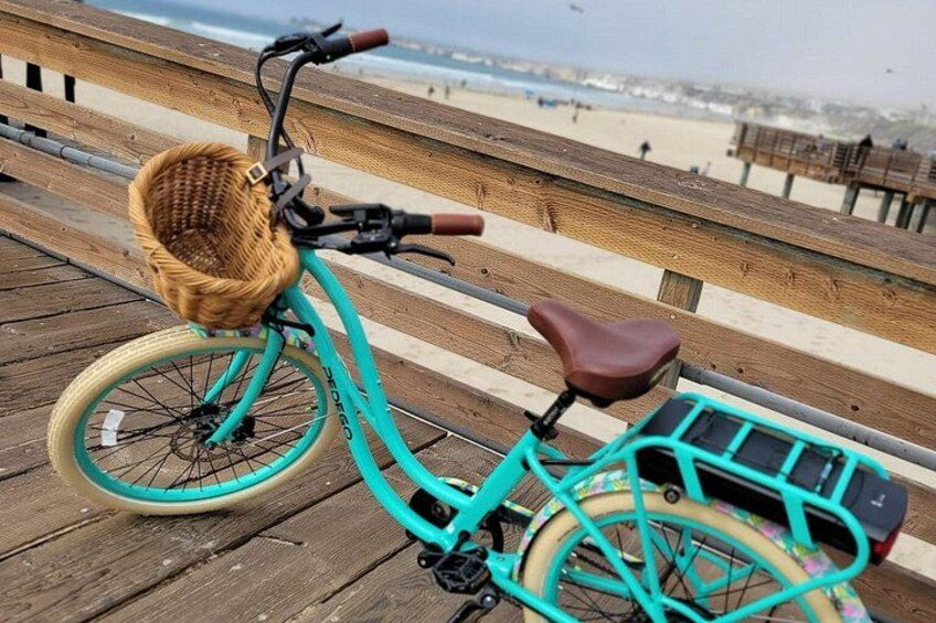 A turquoise bicycle with a wicker basket and a brown seat, parked on a wooden boardwalk with a beach in the background. 4