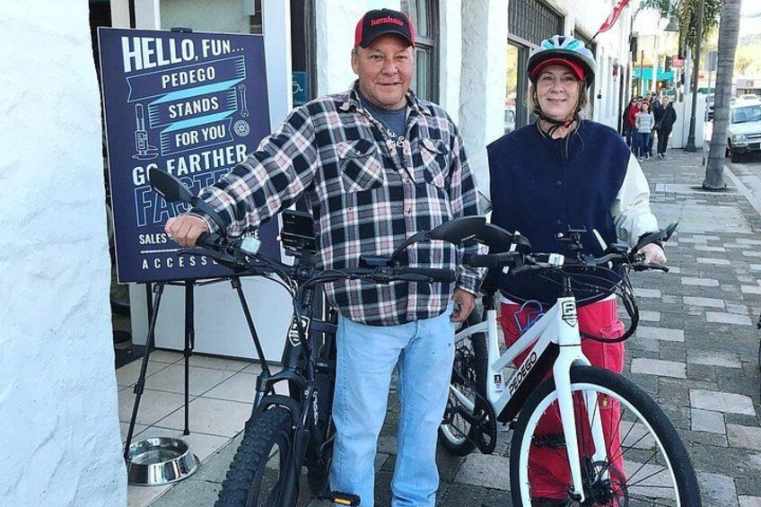Two people standing next to bicycles, a sign with text, and a paved sidewalk. 2