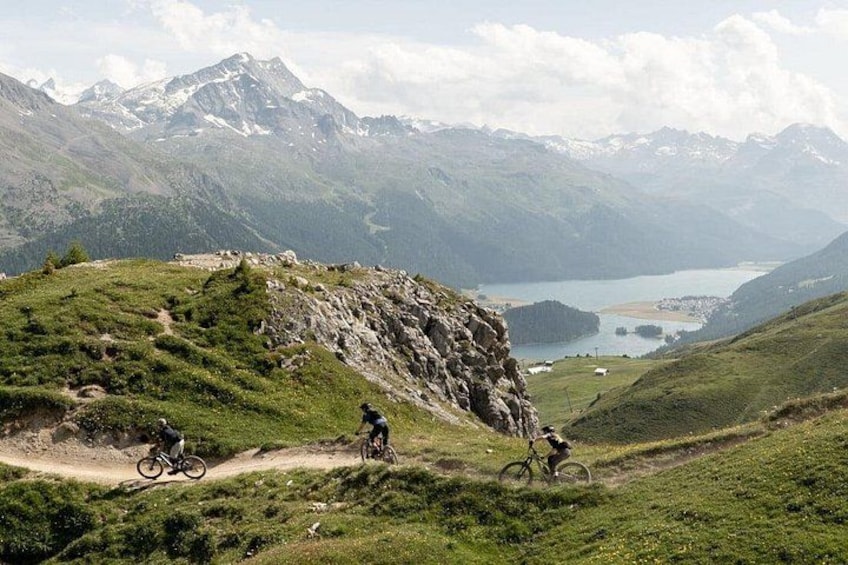 Un percorso per mountain bike con ciclisti, un lago e montagne innevate sullo sfondo. 3