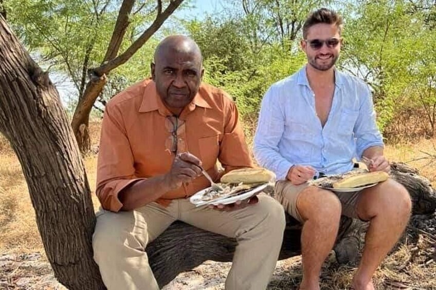 Two people sitting on a tree branch outdoors, one holding a plate with food. 4