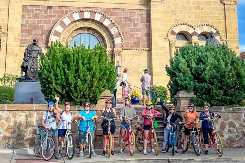 A group of cyclists in front of a historic building with statues and large trees. 1