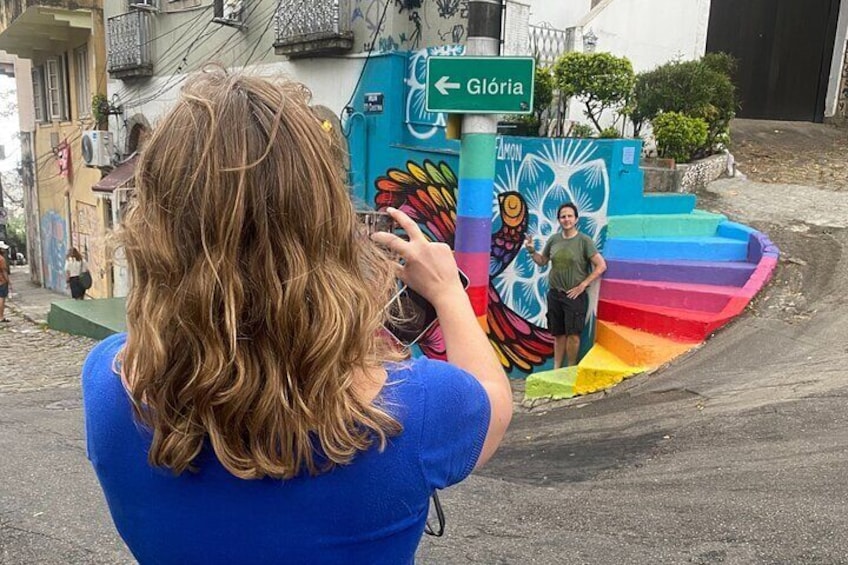 A person taking a photo of a colorful mural with a rainbow staircase and a sign pointing to Glória. 1