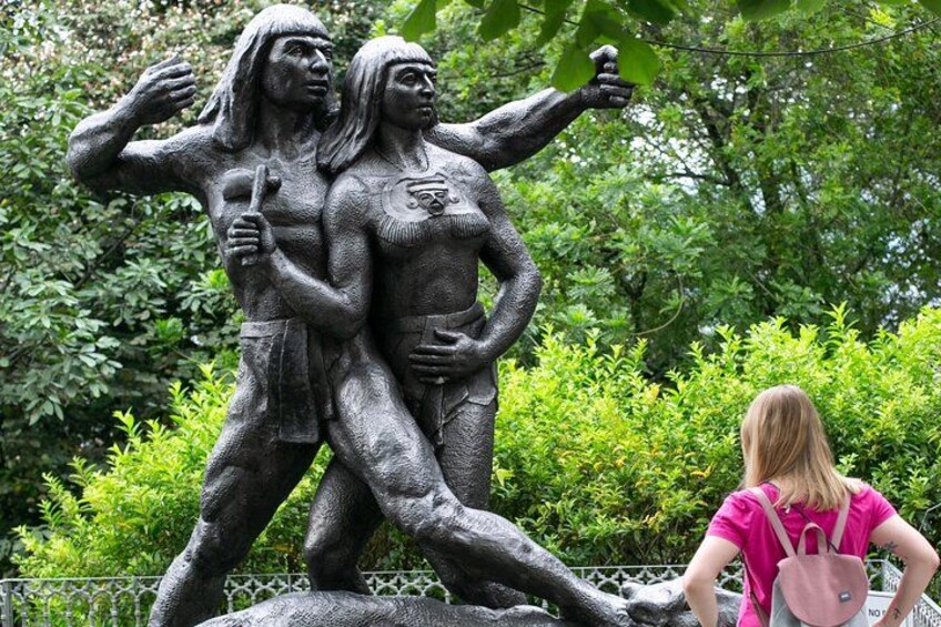 A bronze statue of two figures in a park with greenery in the background. 4