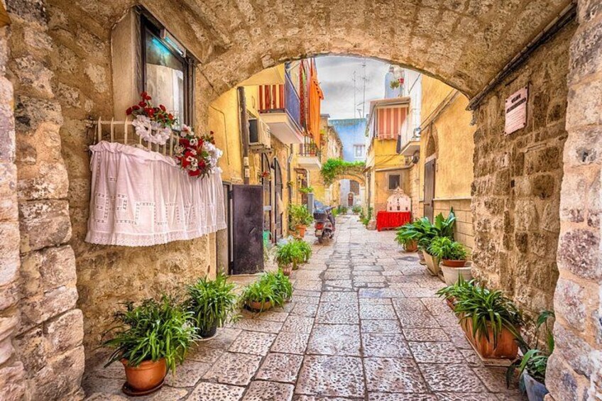 Une ruelle étroite, pavée de pierres, où l’on peut voir du linge suspendu, des plantes en pot et des bâtiments aux couleurs vives. 2