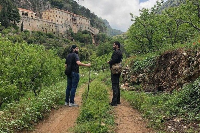 Two people walking on a dirt path surrounded by greenery, with a historic building in the background. 1