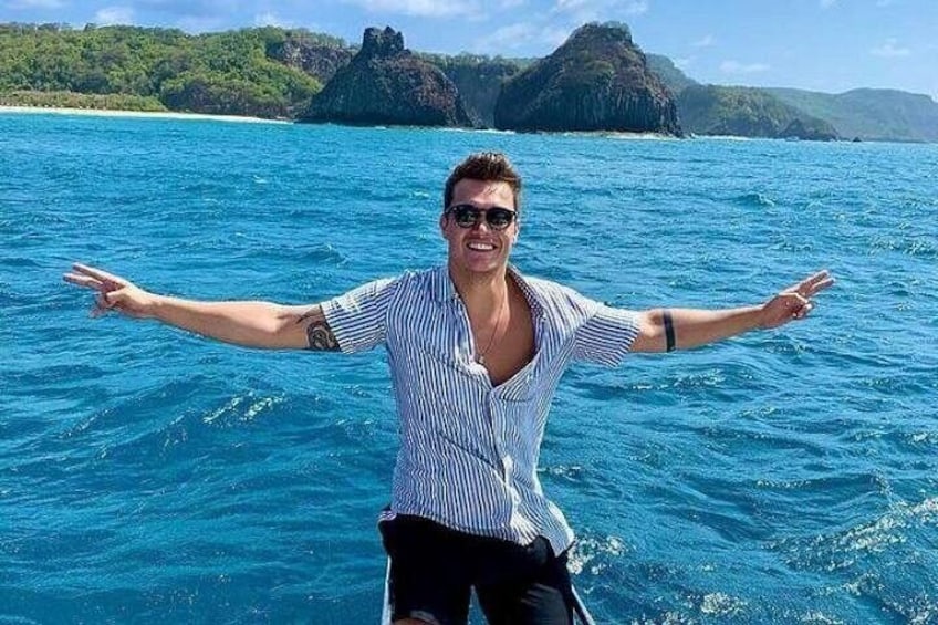 A person standing on a boat with arms outstretched, water, and a distant landmass with vegetation. 1