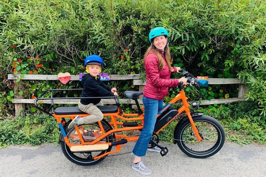 En person på en trehjulig cykel med ett barn sittande bakpå, framför ett trästaket och grönska. 2