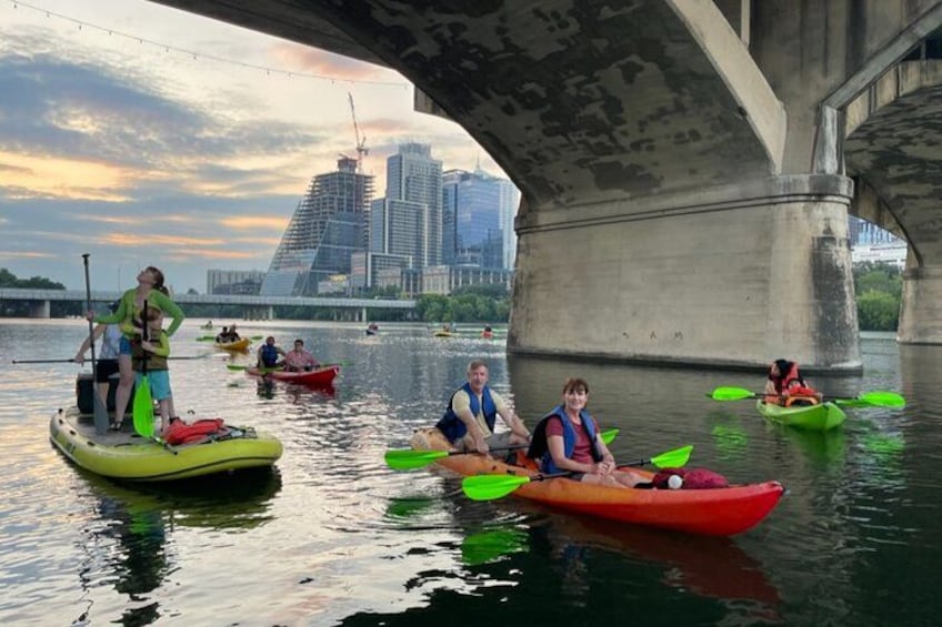 Personas haciendo kayak debajo de un puente, con un horizonte urbano al fondo. 1