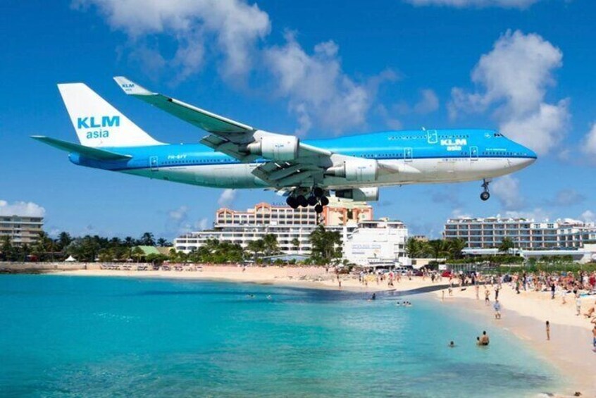 A KLM Asia airplane landing on a beach with clear blue water and buildings in the background. 1