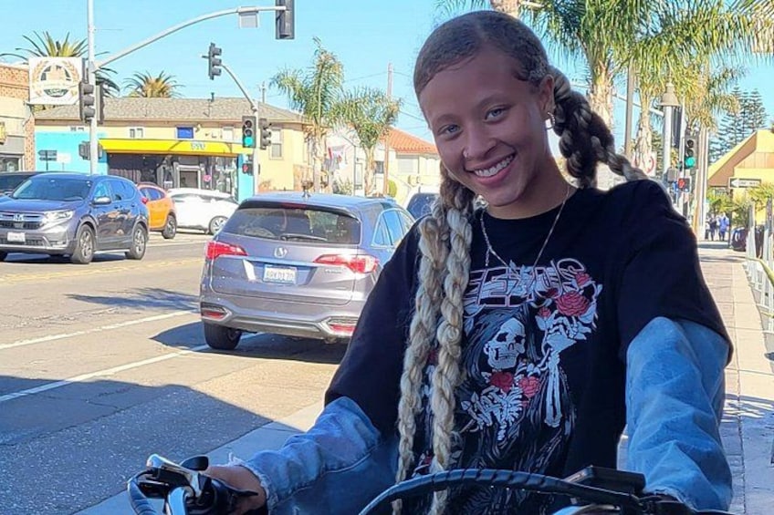A person with braided hair, wearing a black t-shirt with a graphic design, standing on a sidewalk with a bicycle in the foreground. 2