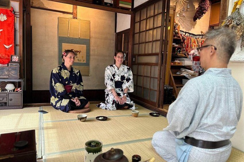 Two individuals in traditional Japanese attire sitting on tatami mats in a room with sliding doors and a shelf with various items. 1
