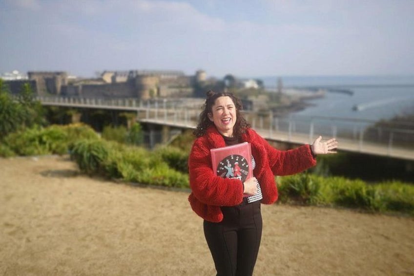 A person in a red jacket holding a pink book, standing on a sandy area with greenery and a bridge in the background. 1