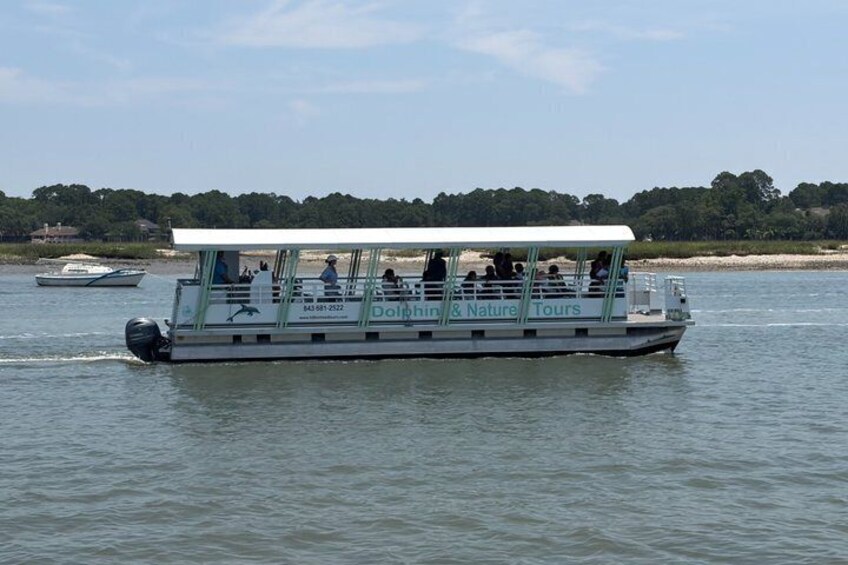 A boat with 'Dolphin & Nature Tours' written on the side, moving on calm water with a shoreline in the background. 2