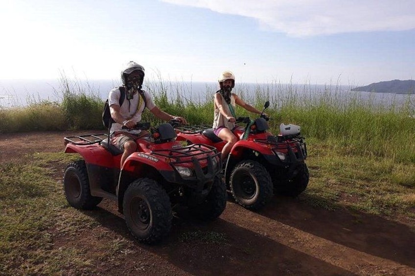 Twee personen op rode quadbikes op een zandpad, met gras en een waterpartij op de achtergrond. 2