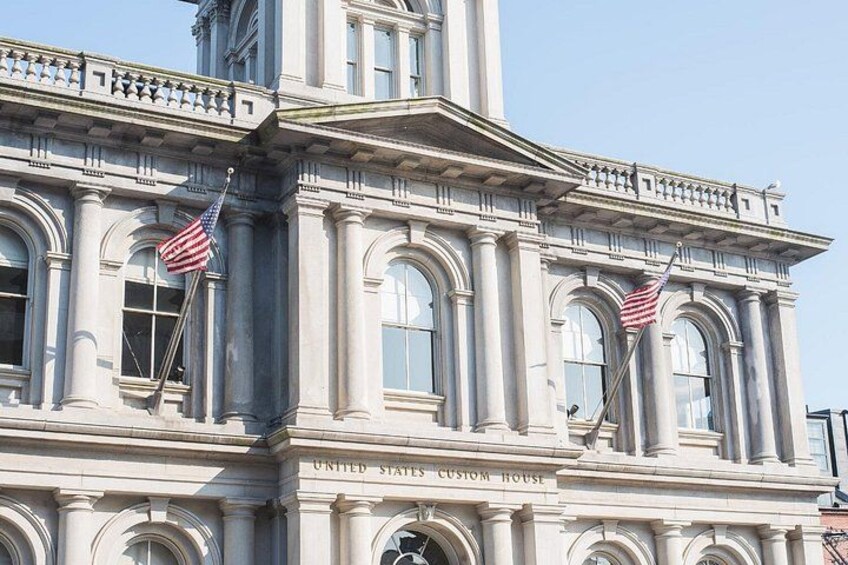 Een historisch gebouw met twee Amerikaanse vlaggen, zuilen en de tekst 'United States Custom House'. 2