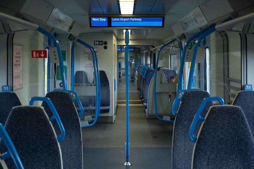 A train interior with blue seats and handrails, a digital display indicating the next stop, and a sign for Luton Airport Parkway. 3