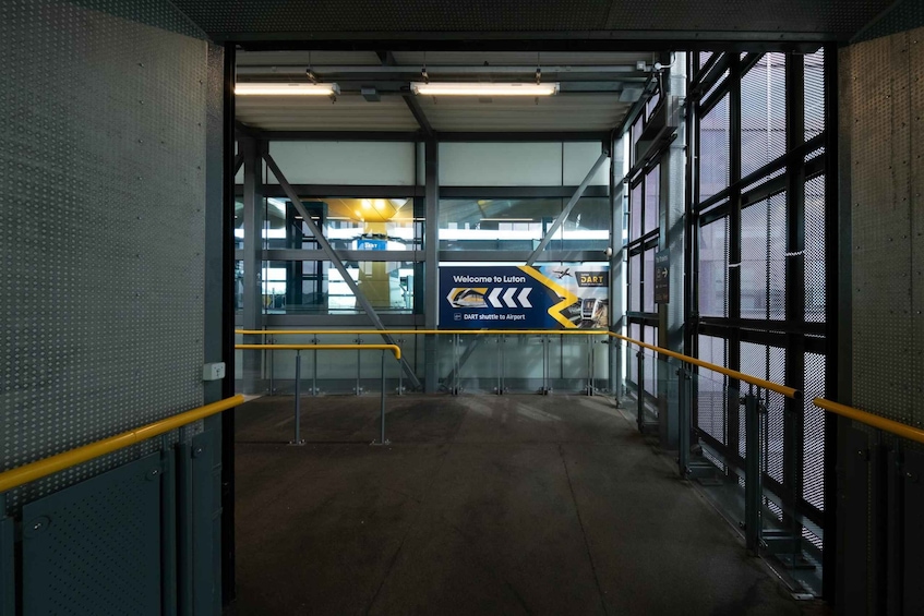 An industrial interior with a sign reading 'Welcome to Luton DART shuttle to Airport' and a yellow directional arrow. 2