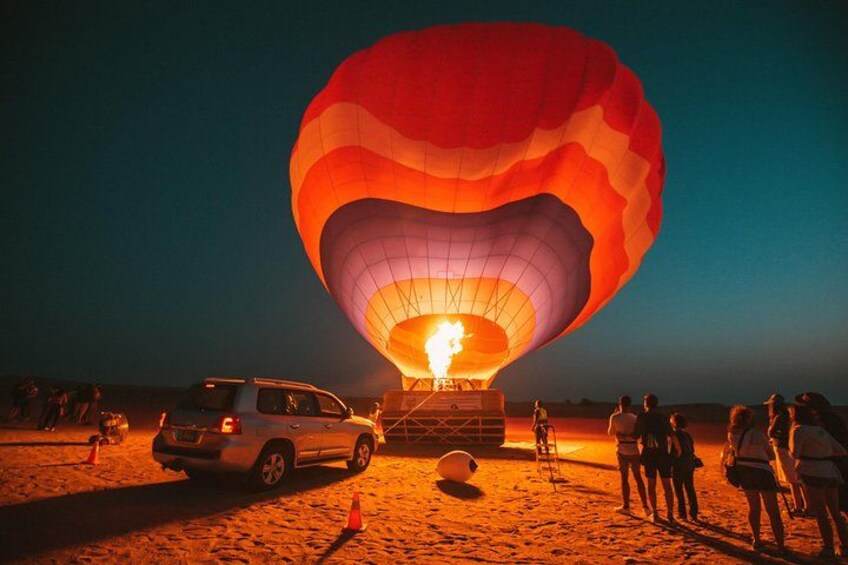Une montgolfière, son brûleur allumé, est garée sur une zone sablonneuse, entourée de personnes et de véhicules. 2