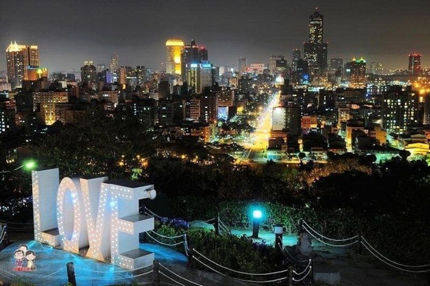 A cityscape at night with illuminated buildings and a large 'LOVE' sign in the foreground. 2