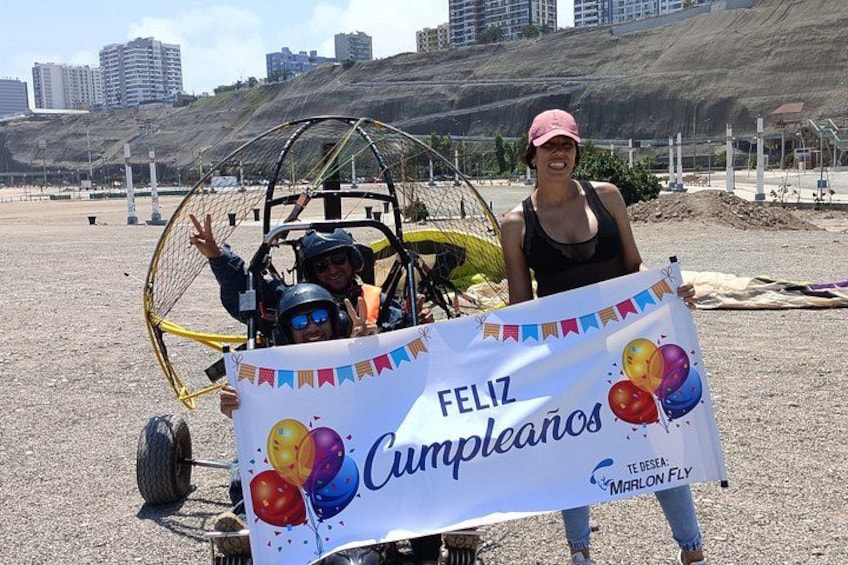 Une personne tenant une banderole d’anniversaire avec des ballons, sur laquelle on peut lire le texte « Feliz Cumpleaños », dans un décor de plage avec une dune et des bâtiments en arrière-plan. 3