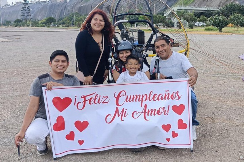 Une famille tenant une banderole d'anniversaire décorée de cœurs et portant le texte « Feliz Cumpleaños Mi Amor ! ». 2