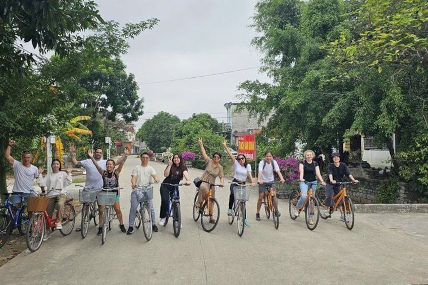 Un grupo de personas en bicicleta, algunas alzando las manos, en un camino pavimentado con árboles y edificios al fondo. 2
