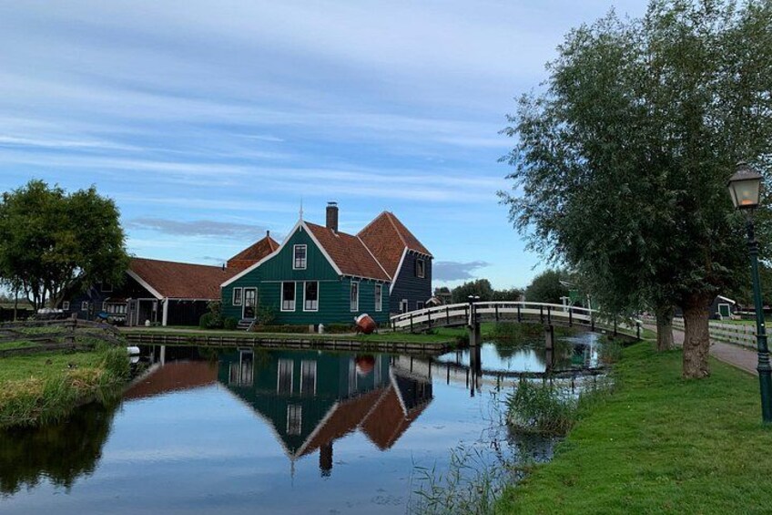 Ett grönt hus med ett rött tak, en liten bro över en stilla vattenyta och ett träd på höger sida. 2