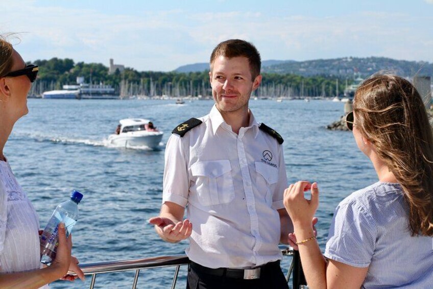 Una persona con uniforme blanco, lleva una etiqueta con su nombre y un distintivo, de pie en un barco, con un cuerpo de agua y otros barcos al fondo. 5