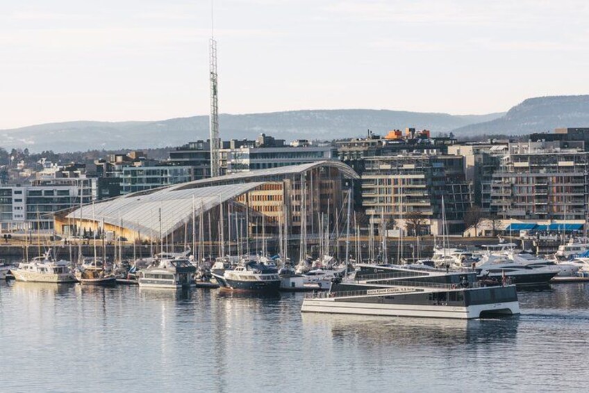 Un puerto deportivo con barcos amarrados, un edificio moderno con techo curvo y un paisaje urbano al fondo. 3