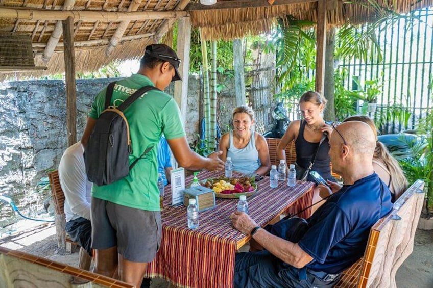 Un grupo de personas cenando en una mesa al aire libre bajo un techo de paja, rodeado de vegetación. 3