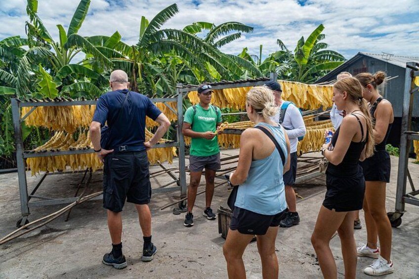 Een groep mensen staat buiten, sommigen dragen casual kleding, met een gebouw en bananenplanten op de achtergrond. 2