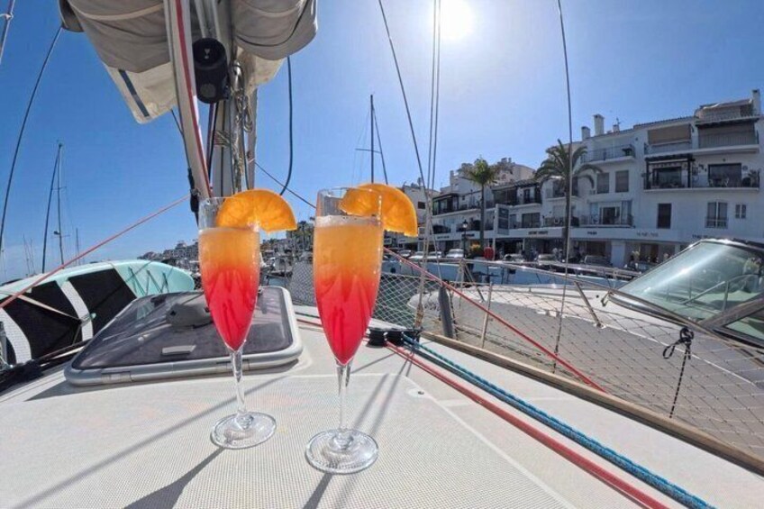 Dos vasos de cóctel rojo con rodajas de naranja sobre la cubierta de un barco, con un puerto deportivo y edificios al fondo. 3