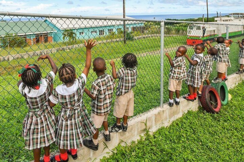 Children in school uniforms standing by a fence, a playground, and a building in the background. 2