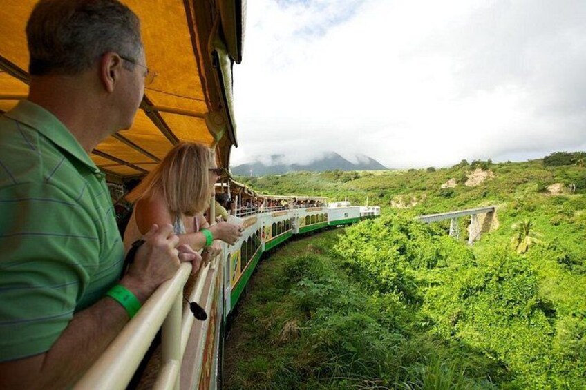 Een treinreis door een weelderig, groen landschap met een brug en een berg op de achtergrond. 1