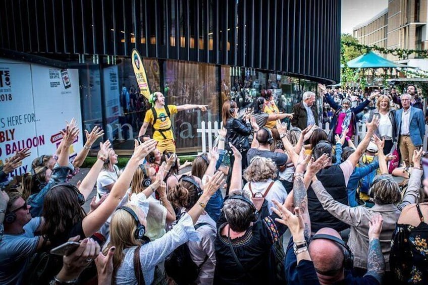 Een menigte verzamelde zich rondom een optredende in een gele outfit, met een gebouw en een bord op de achtergrond. 3