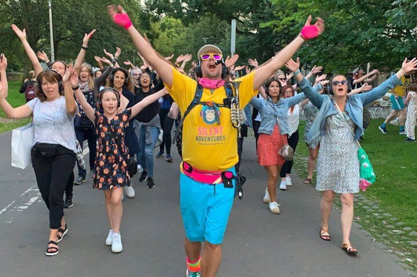 Eine Gruppe von Menschen geht auf einem Pfad, einige mit erhobenen Armen, und eine Person in der Mitte trägt ein gelbes Hemd und eine blaue Shorts. 1