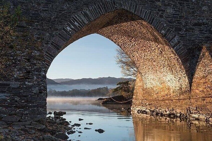 Un puente de piedra arqueado sobre un río tranquilo, con niebla en la distancia y montañas al fondo. 5
