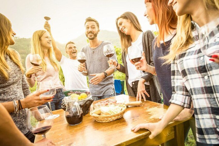 A group of people around a wooden table outdoors, holding wine glasses, with a basket of bread and a bottle of wine visible. 1